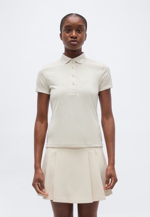 Femme portant un polo beige à manches courtes boutonné et une jupe plissée assortie, debout devant un fond uni gris clair.