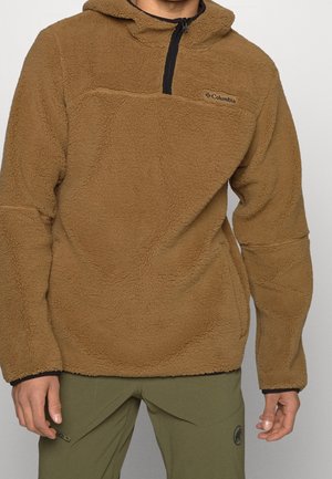 Man die een bruine fleece trui draagt met een zwarte kwartrits en olijfgroene broek, staande tegen een effen achtergrond.