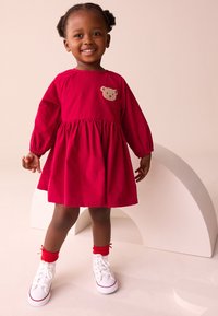 Vestito rosso in cotone con maniche lunghe, gonna arricciata e applicazione orso. Abbinato a scarpe da ginnastica bianche e calzini alla caviglia rossi. Semplice scollo rotondo.