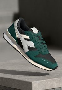 Grønne og svarte athletic sneakers med en overflate av semsket skinn og mesh, med hvite detaljer og såle av gummi, som viser et kontinuerlig gripe mønster.