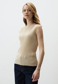 Top beige senza maniche con una texture plissettata, scollatura alta e spacchi laterali, abbinato a jeans blu scuro. Tessuto morbido e liscio.