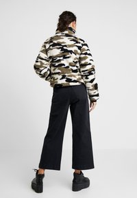 Fleecetröja i beige, svart och olivgrön camouflagemönster; hög krage; mjuk textur. Kombinerad med vida svarta byxor och skor med tjock sula.