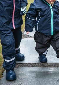 Två barn i marinblå vattentäta jackor och byxor, en med rosa dragkedja, som går på en stenlagd väg. Båda har på sig mörkblå gummistövlar.