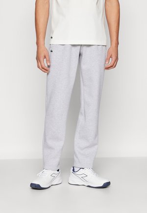 Man draagt lichtgrijze joggingbroek, wit T-shirt en witte sneakers met marineblauwe accenten, staand op een effen lichte achtergrond.