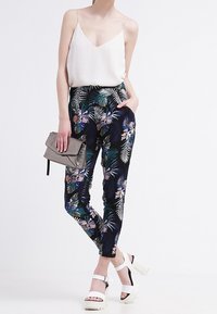 Spodnie w kwiatowy wzór w ciemnym granacie z zielonymi i fioletowymi akcentami, zestawione z białą bluzką na ramiączkach i masywnymi białymi szpilkami.