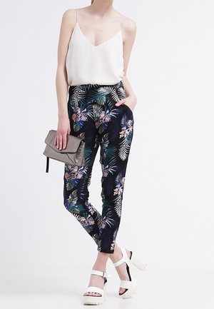 Spodnie w kwiatowy wzór w ciemnym granacie z zielonymi i fioletowymi akcentami, zestawione z białą bluzką na ramiączkach i masywnymi białymi szpilkami.