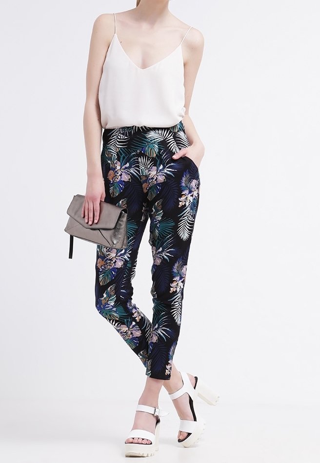 Spodnie w kwiatowy wzór w ciemnym granacie z zielonymi i fioletowymi akcentami, zestawione z białą bluzką na ramiączkach i masywnymi białymi szpilkami.