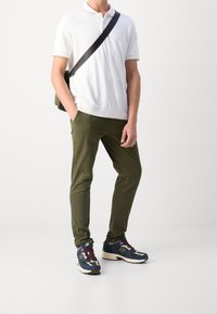 Witte polo met korte mouwen, olijfgroene slim-fit broek en marineblauwe sneakers met meerkleurige accenten; zwarte messengerbag op de schouder.