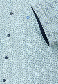 Koszula z kołnierzykiem w jasnoniebieski i zielony geometryczny wzór. Posiada ciemne guziki i czarną kieszeń. Miękka tekstura materiału.