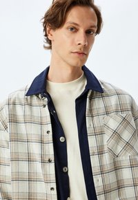 Camisa de botones con un patrón de cuadros beige, cuello azul marino oscuro y un bolsillo en el pecho. Llevada sobre un suéter de cuello redondo color crema.
