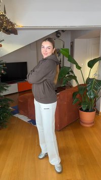 Hanorac maro cu fermoar și șnur, pantaloni albi cu textul „PEGADOR” și adidași argintii. Cadru interior cu plante și un televizor în fundal.