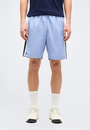 Muž stojaci na jednoduchom pozadí, oblečený v svetlomodrých športových šortkách s bielymi a námorníckymi pruhmi na bokoch, bielych ponožkách a bielych teniskách.