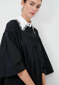 Abito nero con una vestibilità ampia, maniche corte e un colletto bianco con dettagli a taglio. Il tessuto appare liscio e strutturato.