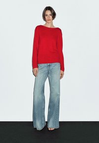 Ein Model steht vor einem schlichten weißen Hintergrund und trägt einen roten Pullover und hellblaue Schlagjeans, die einen lässigen Mode-Look präsentieren.