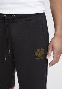 Shorts de algodón negros con cinturilla elástica, cordón, bolsillos laterales y detalle de logo dorado. Textura suave con un diseño casual.