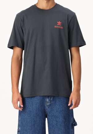 Mand iført mørkegrå Adidas T-shirt med lille rødt logo på brystet og løse blå denimbukser mod hvid baggrund.