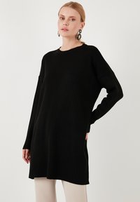 Vestito in maglia nera a coste con maniche lunghe, scollo rotondo e vestibilità ampia, abbinato a pantaloni di colore neutro. Texture in maglia liscia.