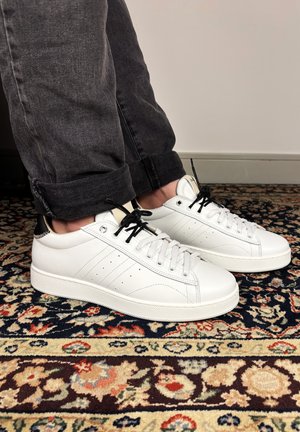 Person wearing black rolled-up pants and white sneakers with black laces standing on patterned multicolor carpet.