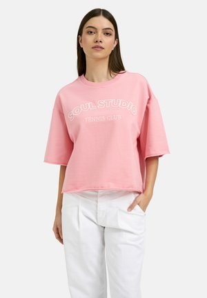 Frau trägt ein pinkes oversized T-Shirt mit dem Text "Soul Studio Tennis Club" und weiße hochtaillierte Hosen, steht mit einer Hand in der Tasche.