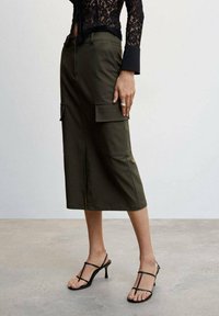 Jupe midi vert olive avec deux poches cargo latérales, coutures apparentes et coupe droite. Associée à des talons noirs à lanières.