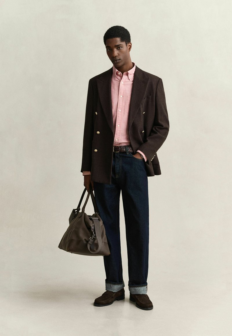 Hombre que lleva un blazer marrón de doble botonadura, camisa rosa abotonada, vaqueros oscuros remangados, zapatos marrones, sosteniendo un bolso de cuero marrón con una mano.