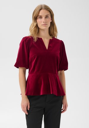 Femme portant un top en velours rouge à manches courtes avec un col en V et un ourlet péplum, associé à un pantalon noir, debout devant un fond uni.