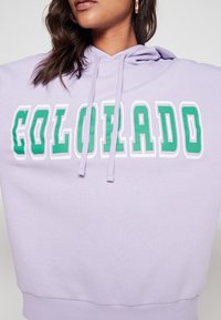Donna che indossa un felpa leggera di colore viola con la scritta "COLORADO" in verde e bianco sul petto, mostrando parzialmente il viso e i capelli.