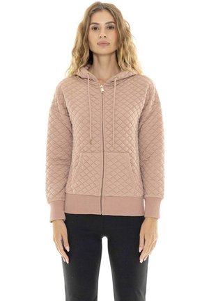 Une femme porte un sweat à capuche matelassé rose poudré, avec une fermeture éclair et des cordons, ainsi qu’un pantalon noir sur fond blanc.