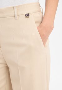 Beige herringbone-patterned pants with a side pocket and a hand partially inserted, featuring a small leopard print belt loop detail.