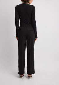 Schwarzes Langarmshirt mit taillierter Passform, kombiniert mit lockeren schwarzen Hosen mit Seitentaschen und knöchellangen Säumen über Absatzschuhen.