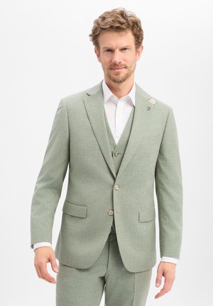 Mann trägt hellgrünen Dreiteiler und weißes Hemd, steht vor schlichtem hellem Hintergrund, blickt nach vorne.