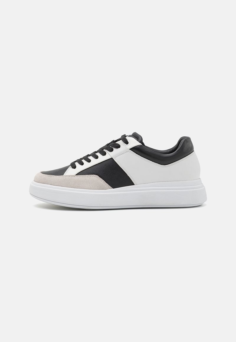 Sneaker bas profil avec des panneaux noirs, blancs et beiges, des lacets noirs et une épaisse semelle blanche, photographié sur un fond gris clair uni.