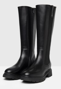 Botas negras hasta la rodilla hechas de cuero suave, con una cremallera lateral y paneles elásticos para mayor flexibilidad, con una suela gruesa y con dibujo.