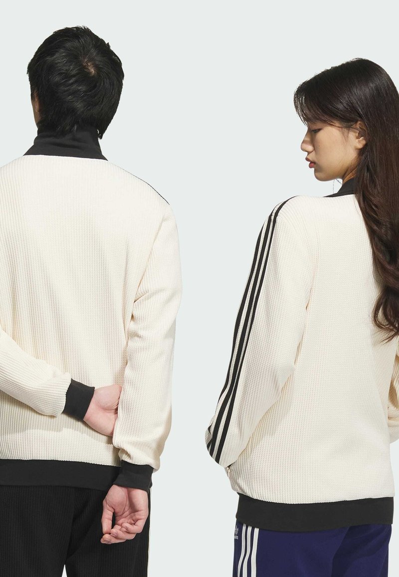Cremefarvet ribstrikket pulloverjakke med sorte accenter og tre striber ned ad ærmerne. Har høj hals og lange ærmer.