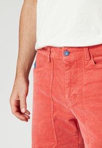 Röd kordshorts med en blå knappstängning, framfickor och ribbad textur. Designen är åtsittande vid midjan.