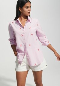Light pink striped shirt with red lobster embroidery, long sleeves rolled up, collared design, and a relaxed fit. Worn with white shorts.