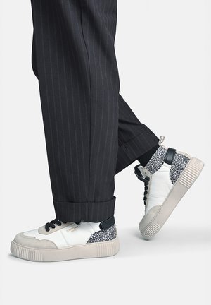 Person iført sorte bukser med striber og hvide høje sneakers med sort-hvide leopardprint-accenter.