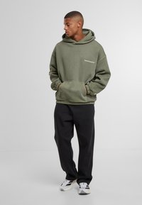 Joven con una sudadera verde oliva, pantalones negros y zapatillas blancas, de pie con las manos en los bolsillos de la sudadera mirando de lado.