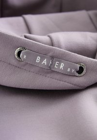 Etichetta in tessuto lilla con il nome del marchio "BAIER" in lettere bianche, attaccata a un capo textured con un occhiello in argento.