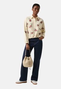 Kremowy sweter z kwiatowym nadrukiem, duży kołnierz, elastyczne mankiety, w połączeniu z ciemnymi dzwonowatymi spodniami z denimu i beżową torbą typu bucket. Białe trampki.