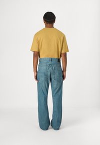 Camiseta de algodón amarilla de manga corta, combinada con jeans de mezclilla azul claro con bolsillos traseros y un ajuste relajado. Vista completa desde atrás.