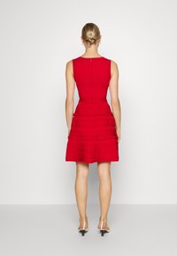 Robe sans manches rouge avec un corsage ajusté, un ourlet à volants, un tissu texturé et une fermeture éclair au dos. Portée avec des sandales à talons blanches.