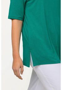 Maglietta verde a maniche corte realizzata in materiale morbido con aperture laterali, caratterizzata da una vestibilità ampia e dettagli di cucitura visibili, abbinata a pantaloni bianchi.