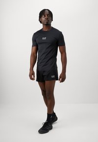 Svart treningst-skjorte og shorts med EA7-logo, laget av glatt syntetisk materiale. Kombinert med svarte sportsko og sokker.