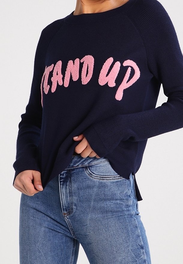Marinblå tröja med rosa texturerad "STAND UP"-bokstäver, ribbade ärmslut och sprund i sidorna, med ljusblå jeans i denim.