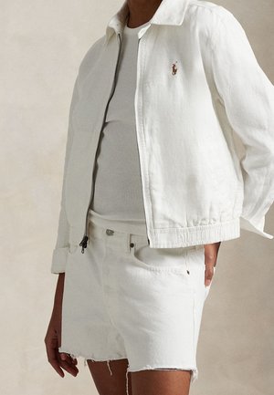 Personne portant une veste blanche zippée sur un top blanc côtelé associée à un short en denim blanc avec des ourlets effilochés.