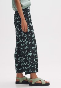 Zwarte wijde broek met een teal abstract patroon, gemaakt van zachte stof. Gecombineerd met groene sandalen met banden en een platte zool.