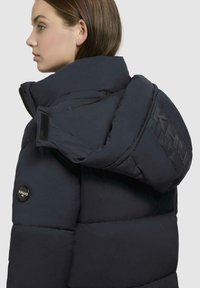 Pufferjakke i mørk marineblå, med høj krave, store skuldre, synlige sømme, logo-patch og lynlåslommer på siden.