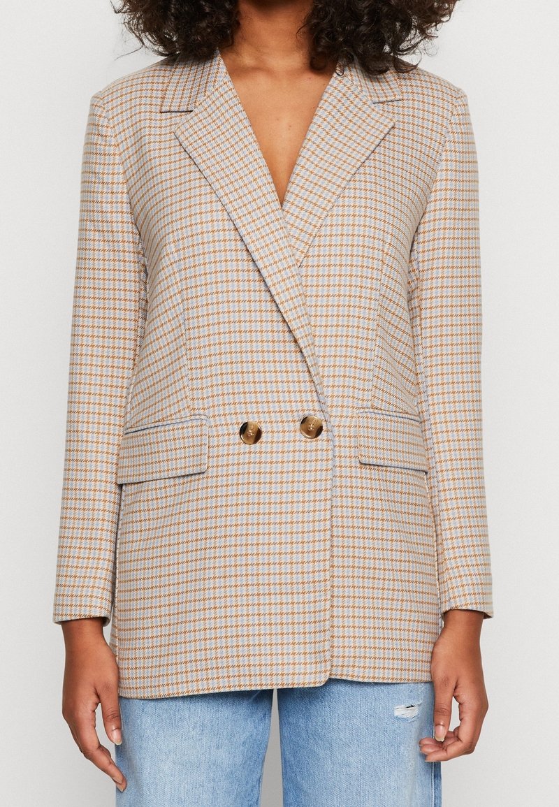Femme portant un blazer à double boutonnage beige et bleu clair à carreaux avec poches à rabat et un jean bleu clair déchiré.