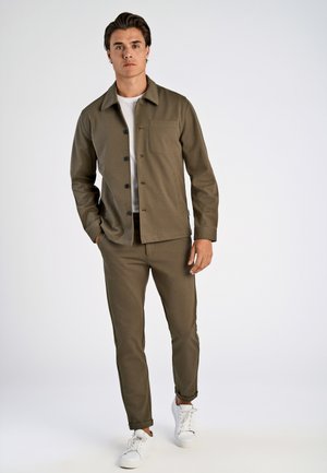 Chaqueta color oliva con botones negros, un bolsillo en el pecho, combinada con pantalones entallados a juego y zapatillas blancas. Tela suave, diseño casual.
